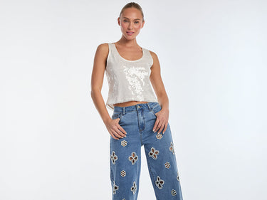 A person wearing a silver sequin sleeveless top and blue wide-leg jeans with floral embroidery stands confidently against a plain background.