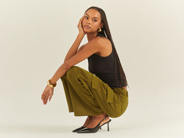 A person with long braided hair wearing a black sleeveless top, wide-leg green pants, and black heeled shoes squats on the floor against a light grey background, with one hand touching their face and the other resting on the floor.