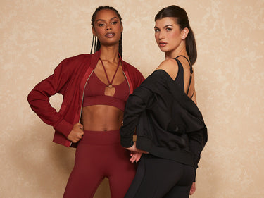 Two women stand confidently in athletic wear against a neutral background. One wears a burgundy set with a matching jacket, the other in a black set with a matching jacket draped over her shoulders.