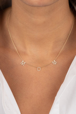 Close-up of a person wearing a white shirt and the Diamond Pave Mom Necklace 14K by By Adina Eden, featuring an "M," an open circle, and another "M" pendant. The shirt is unbuttoned at the top, revealing part of the neck and upper chest.