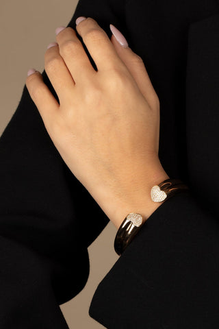 A person wearing a black coat is seen with their left hand resting on their right forearm, showcasing the stylish Pave Heart Open Claw Bangle Bracelet by Adina Eden. The bracelet features two shimmering heart-shaped embellishments adorned with clear CZ stones and is plated in 18K gold. The background is a neutral beige color.