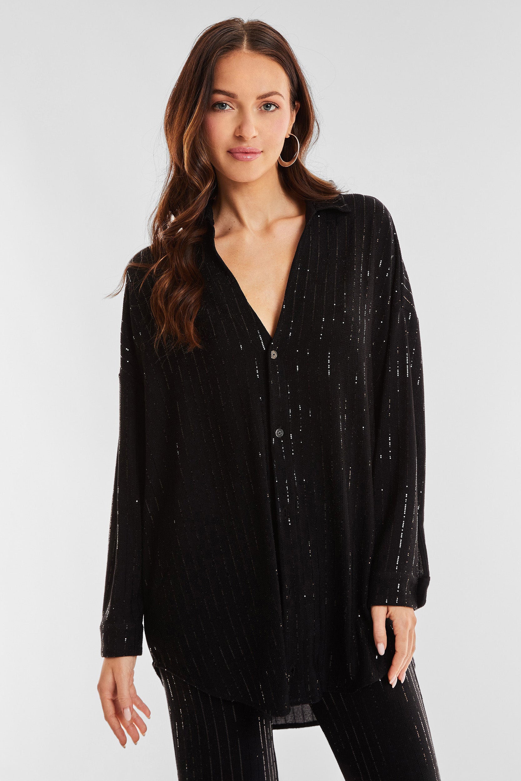 A person with long, wavy hair is wearing an Amalfi Embellished Shirt - Black, a long-sleeved button-up featuring subtle vertical shimmering stripes. They are posing against a plain light gray background, looking directly at the camera with a neutral expression.