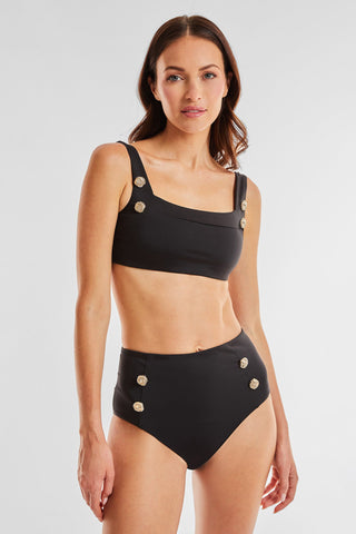 A woman stands against a plain backdrop, wearing the Mykonos Bottom - Black, part of a two-piece swimsuit adorned with metallic buttons. The top features thick straps and the high-waisted bottom offers tummy control, creating a slimming effect. She has long, wavy hair and a neutral expression.