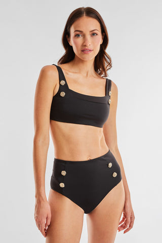 A woman with long brown hair stands against a plain white background, wearing the black Mykonos Top by Noli Yoga, which features gold decorative buttons on the wide shoulder straps and is paired with high-waisted bottoms for a slimming effect.