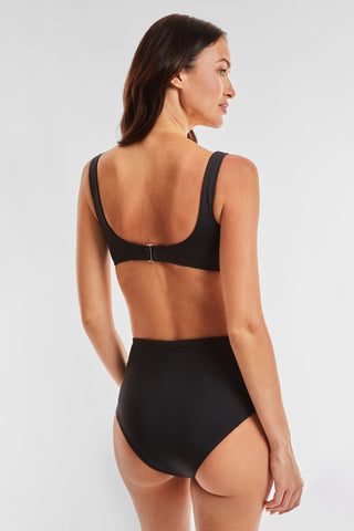 A woman models the Mykonos Top - Black, part of a bikini swimwear set. She stands with her back turned, looking over her shoulder. The high-waisted bottom and top with broad straps offer a slimming effect, and a clasp at the back completes the swimsuit's sleek design against the plain white background.