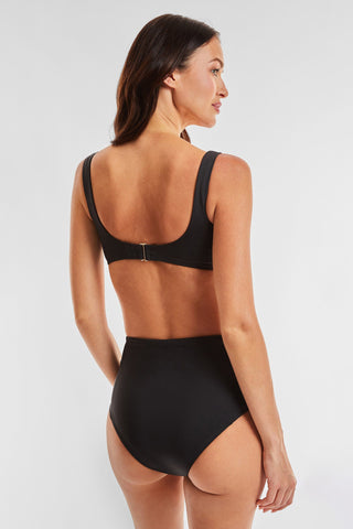 A woman in a black bikini stands with her back to the camera, highlighting the Mykonos Bottom - Black, known for its high-waisted tummy control design. The swimsuit has a top featuring a clasp closure, all against a simple white background.