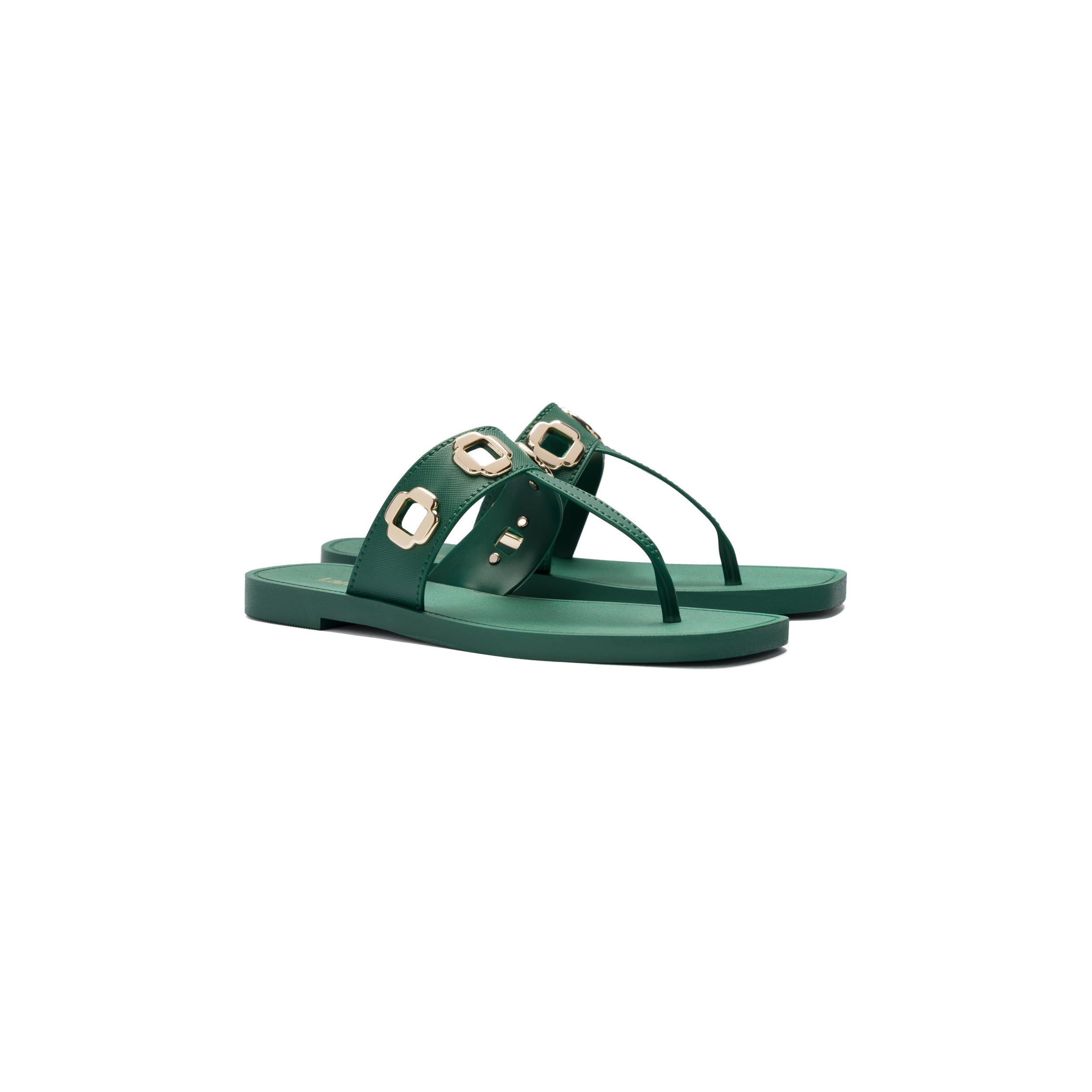 The Milan S In Emerald PVC sandals are a pair of stylish green shoes featuring a flat sole and backless design. These Larroudé bestsellers are adorned with golden hexagonal studs on the t-strap, providing both security and a decorative touch. The thong-style strap runs between the toes.