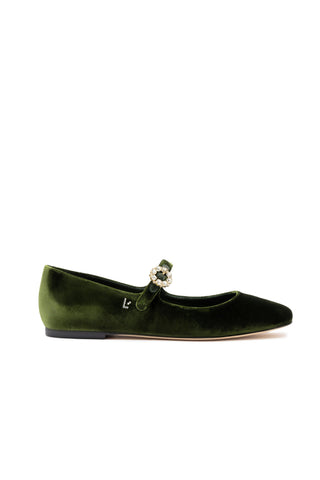 The Blair Ballet Flat in Bosco Velvet is a single green velvet Mary Jane shoe that features an adjustable ankle fastening adorned with a decorative rhinestone buckle. The shoe showcases a small "l" detail on the side, has a slightly pointed toe, a flat heel, and includes memory foam cushioning for added comfort.