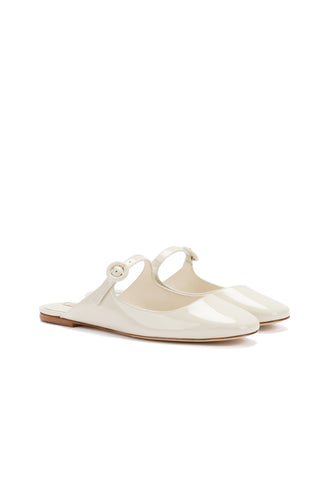 The Blair Flat Mule in ivory patent leather is a pair of glossy slip-on shoes with a low heel and a thin ankle strap featuring a small buckle. This adorable, Mary Jane-inspired style boasts a sleek, minimalist design with closed, rounded toes and a smooth, shiny finish.