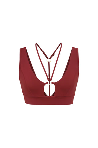 The Esme Bra - Merlot is a red bralette featuring a deep V-neckline and a unique strappy design with an adjustable ring detail in the center. It has wide shoulder straps, a front cut-out, and is made of smooth fabric, perfect for low to medium impact workouts.