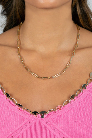 A woman wearing the Large Paperclip Link Necklace by Adina Eden and a pink textured top with decorative metallic straps. The top has a detailed hem and metallic, circular accents lining the straps. The focus is on the versatile accessory necklace and the upper portion of the top.