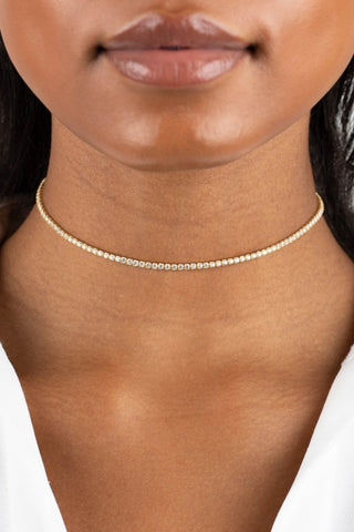 A close-up of a person wearing a delicate Thin Tennis Choker by By Adina Eden, featuring small, evenly spaced round beads. The person has medium-dark skin and is dressed in a white shirt with an open collar. Only the lower part of their face and neck are visible against a plain white background.