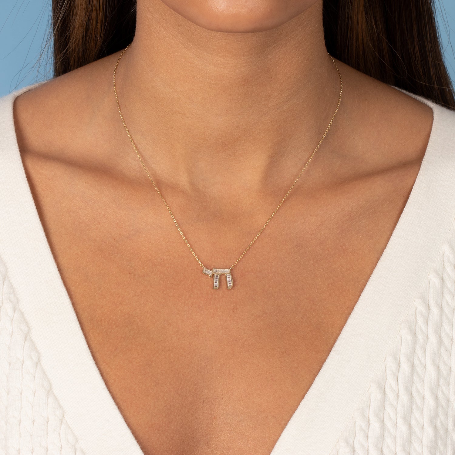 A close-up showcases a person wearing the delicately crafted Baguette Chai Pendant Necklace by Adina Eden. This exquisite piece features three vertical bars, each adorned with sparkling clear baguette CZ stones, all set in thin, yellow gold. The individual is dressed in a white v-neck knit sweater, and their straight brown hair cascades slightly against a light blue background.