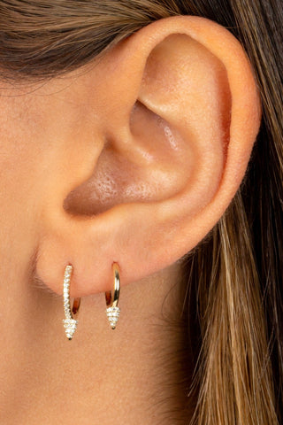 A close-up of an ear adorned with two huggie earrings. The first earring boasts a plain 14K gold finish, while the second one, named "Diamond Point Huggie Earring 14K by By Adina Eden," showcases a gold finish embellished with a row of small pavé diamonds. Strands of light brown hair partially obscure the background.