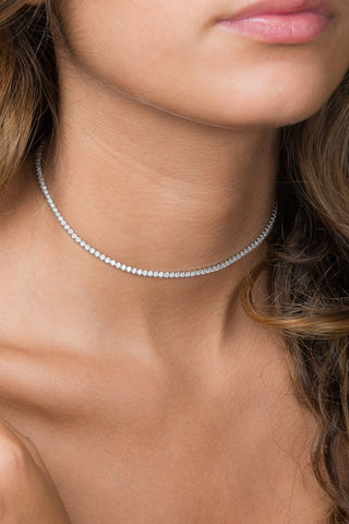 A close-up image of a person wearing the Thin Tennis Choker by By Adina Eden, a delicate, gold-plated sterling silver necklace adorned with dazzling cubic zirconia stones. The person's shoulder-length, wavy hair cascades around their bare shoulder, emphasizing the elegance of the necklace against their skin.