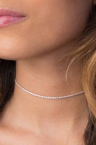 A close-up of a woman's lower face and neck, showcasing her wearing the elegant Thin Tennis Choker by Adina Eden, adorned with dazzling CZ stones. Her lips have a glossy pink tint, and her wavy brown hair falls over her shoulders. The image exudes elegance and minimalistic style.