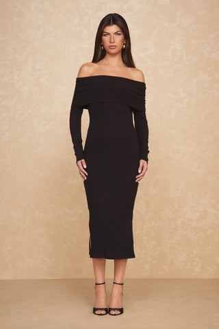 A woman stands against a beige textured background, wearing the ultra-soft Amora Dress - Black, an off-the-shoulder midi dress with long sleeves. She is also wearing black high-heeled sandals and gold earrings. Her hair is styled straight, and she has a neutral expression.