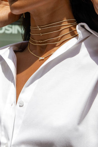 A person wearing a white button-up shirt showcases their neck adorned with multiple layered Thin Tennis Chokers by By Adina Eden, each featuring small, delicate pendants.