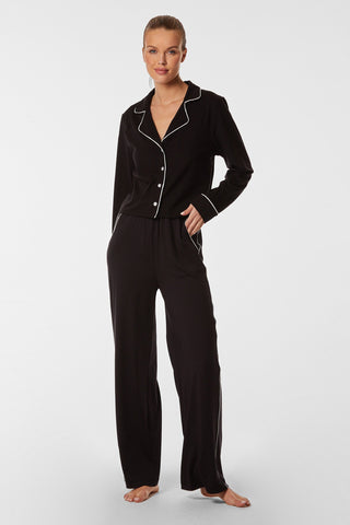 A woman stands against a white background wearing the Black Pajama Soft Long Pant set with white piping. The soft, high-waisted pants perfectly complement the long-sleeve button-up top with a collar. She has one hand in her pocket and is barefoot, ready to lounge around comfortably.