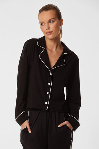 A woman with light brown hair tied back is wearing the Black Pajama Soft Long Sleeve Shirt, featuring white piping and buttons. She has her hands in her pockets and is looking to her left with a relaxed expression against a plain white background.