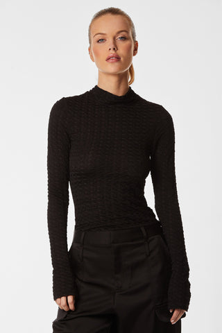 A person with light-colored hair tied back is wearing the Florence Sheer Turtleneck in black and high-waisted black pants. They are standing against a plain white background, looking directly at the camera with a neutral expression.