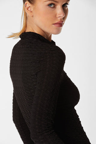 A person with light-colored hair pulled back is gazing over their shoulder at the camera. They are wearing a Florence Sheer Turtleneck - Black, featuring a textured design. The background is plain and white.