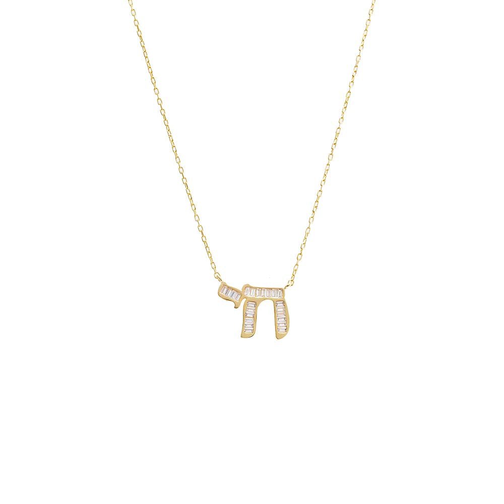 The Baguette Chai Pendant Necklace by Adina Eden is a delicate gold necklace featuring the Hebrew word "Chai," which symbolizes life. This elegant piece is adorned with clear baguette CZ stones that are arranged in a rectangular pattern along the letters. The necklace boasts a thin and minimalistic chain made of sterling silver plated with gold.