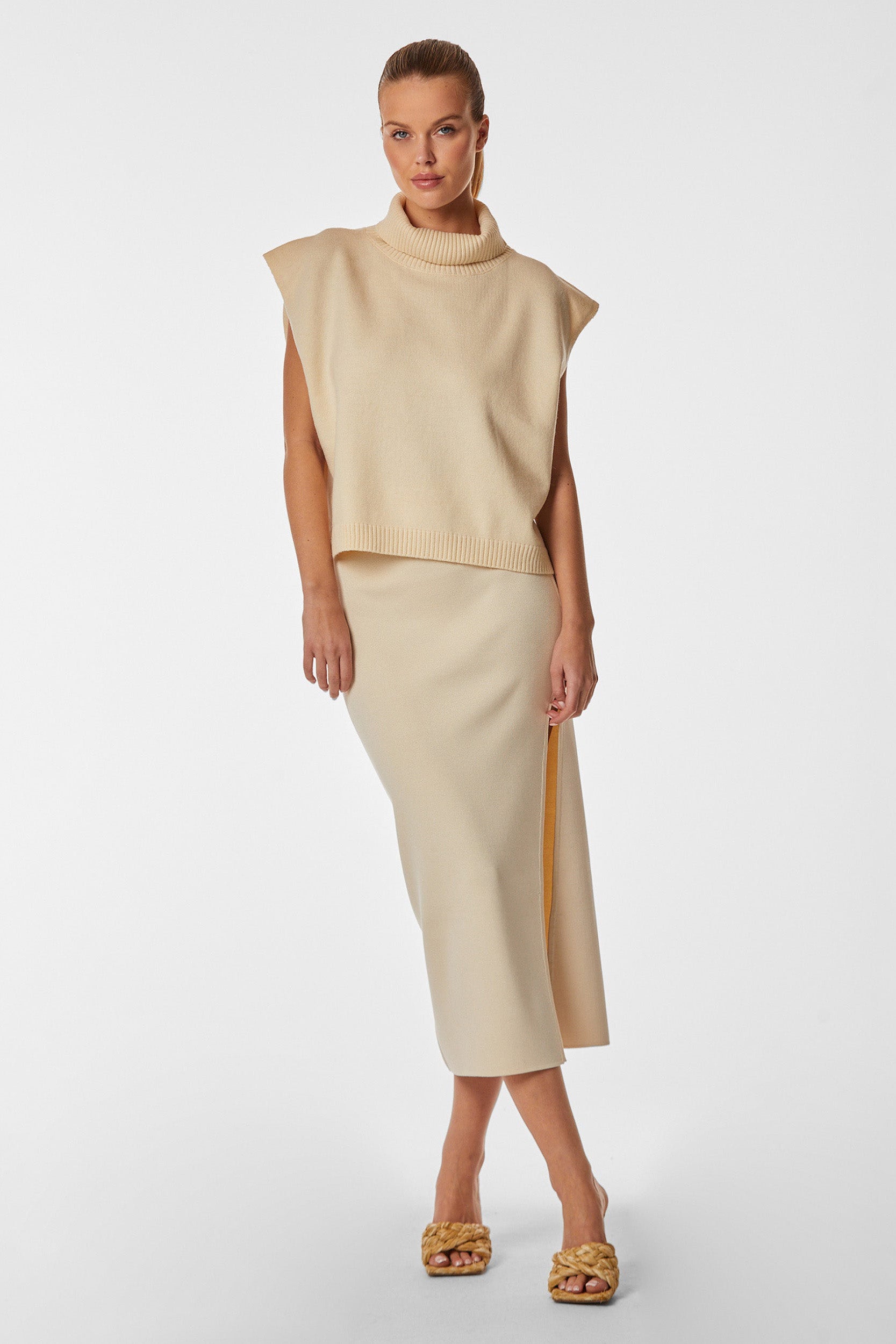 A person is standing against a plain white background wearing a beige sleeveless turtleneck sweater paired with the Hamptons Midi Skirt - Pearl, featuring a front slit and slim-fitted silhouette. They are also wearing yellow textured sandals. The person's hair is pulled back in a sleek style, and they have a neutral expression.