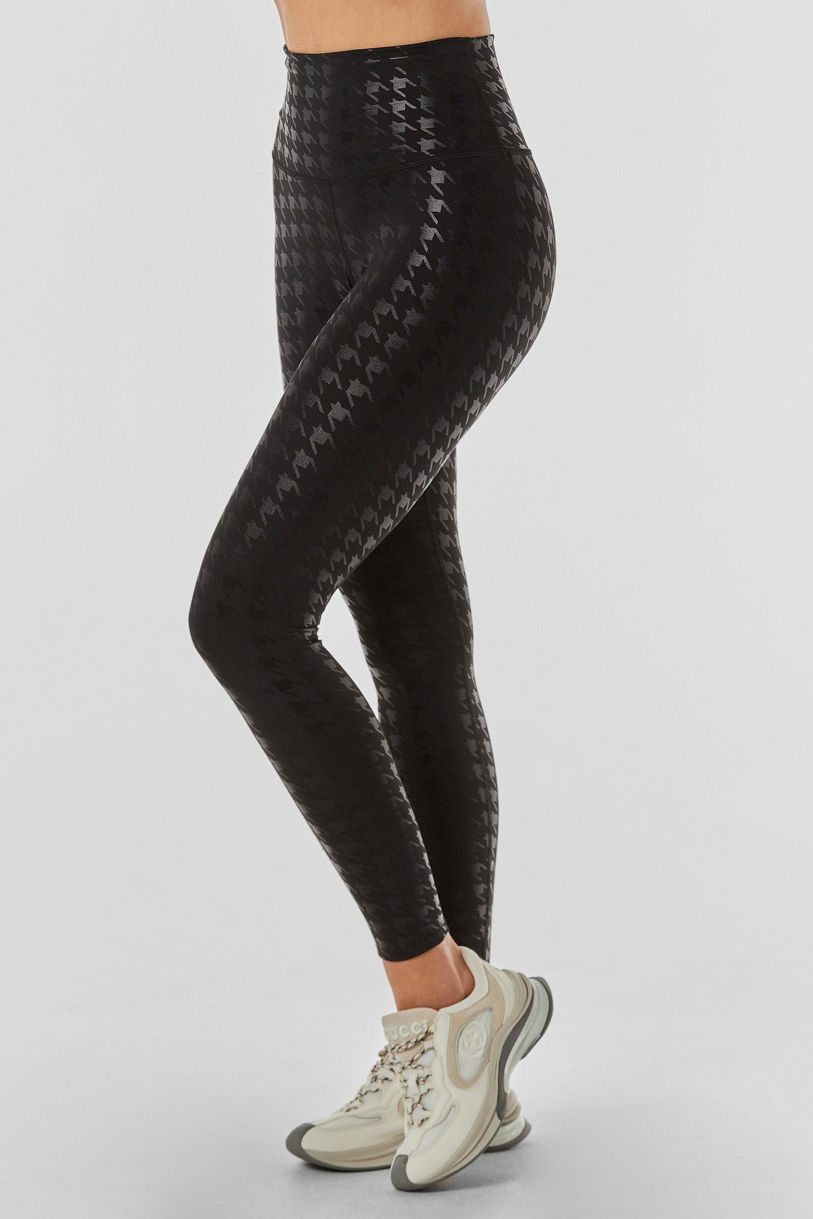A person is wearing the Houndstooth High-Waisted Legging - Black, crafted from supportive performance fabric. The black leggings showcase a glossy houndstooth pattern and include a four-way stretch waistband. They are paired with white sneakers featuring light green and grey accents. The plain white background highlights only their lower body.