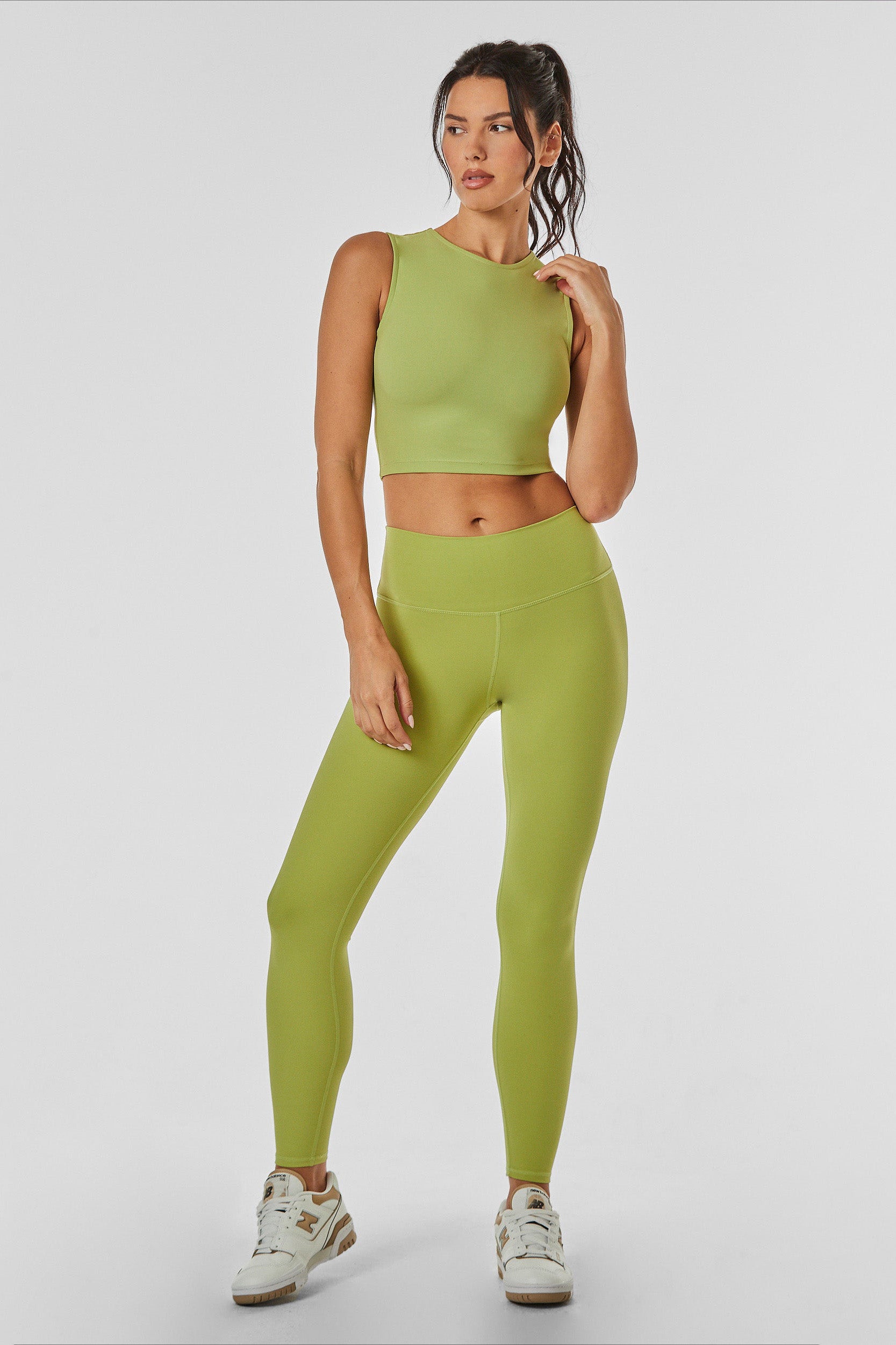 A woman stands confidently against a plain background, wearing a matching green athletic outfit consisting of a sleeveless crop top and the Limitless Legging in Matcha. She has her hair tied back in a ponytail and is wearing white and green sneakers.