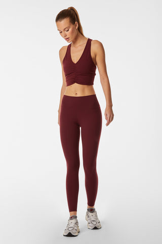 A woman stands against a plain white background, wearing the Limitless Legging - Pinot, a matching burgundy workout set that includes a sleeveless, V-neck crop top and high-waisted, moisture-wicking leggings. Her long, straight hair is pulled back in a ponytail, and she completes her look with white sneakers. She looks downward.