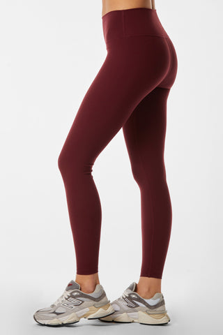 A person is seen from the side, wearing Limitless Legging in Pinot, which are maroon high-waisted sculpting leggings made of moisture-wicking fabric. They are also sporting gray sneakers with white and beige accents. The background is plain white.