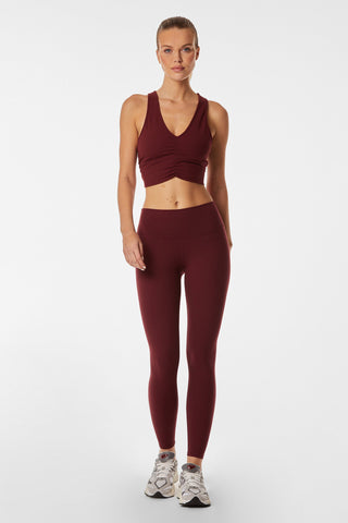 A woman stands against a plain background wearing a burgundy sports bra and the Limitless Legging in Pinot. She has her hair tied back and is wearing white athletic sneakers. The moisture-wicking fabric keeps her comfortable as she faces forward, looking directly at the camera.