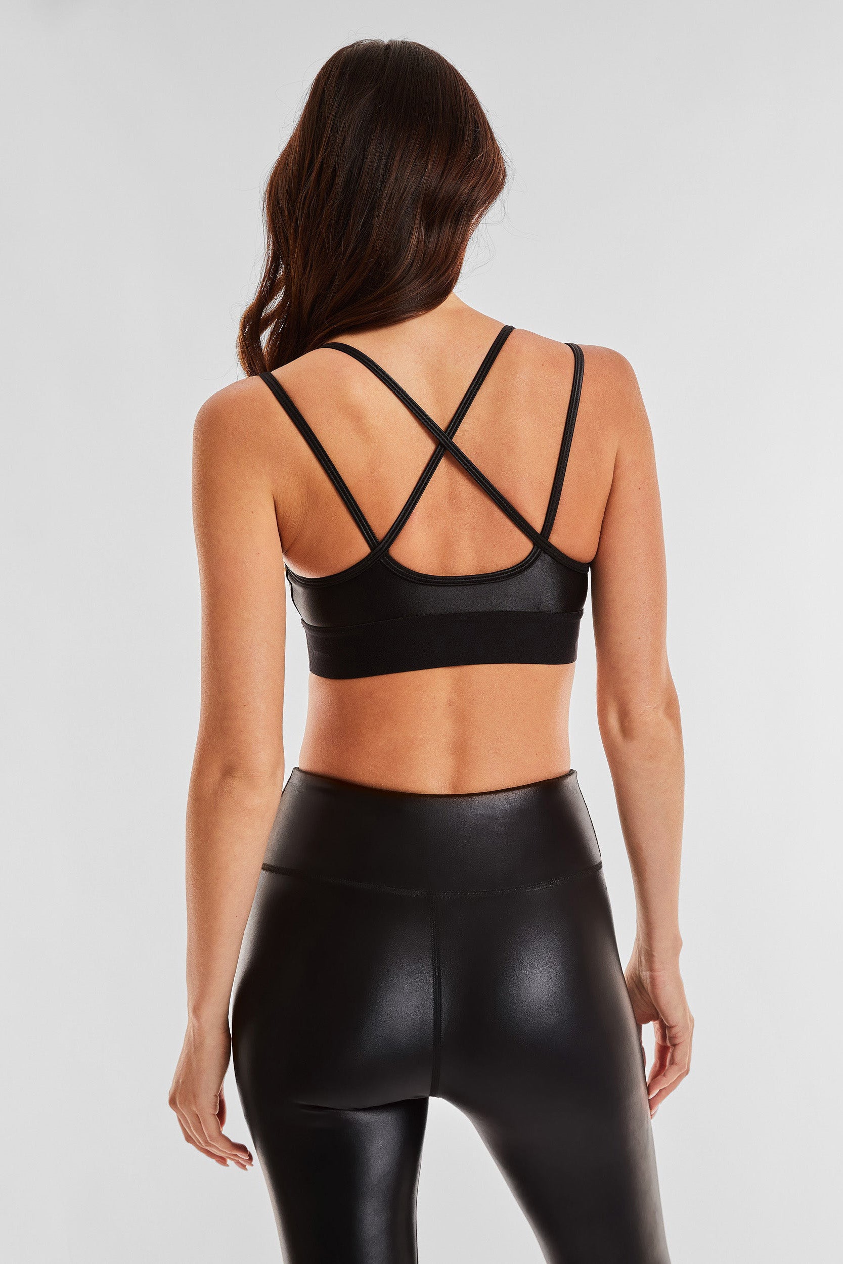 A woman with long wavy hair is shown from behind wearing a black strappy Liquid Jolie Bra - Black Gloss and high-waisted black leggings. The moisture-wicking sports bra features an intricate criss-cross strap design on the back. The background is plain and white.