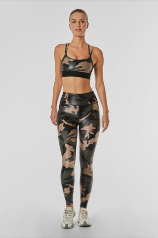 A woman stands against a plain background, wearing a camo-patterned Liquid Jolie Bra with strappy back details and matching leggings. She also has on white sneakers. The outfit is moisture-wicking and designed for fitness activities.