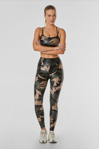 A person stands against a plain background wearing a matching Liquid Jolie Bra - Camo with a strappy back and leggings. They have their arms crossed, and they are also wearing white athletic shoes. Their hair is pulled back.