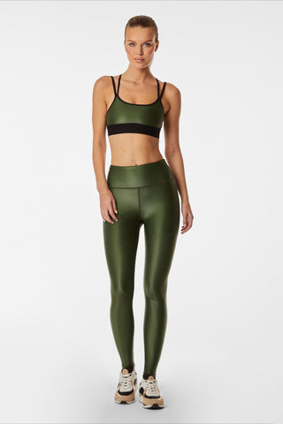 A person standing on a plain white background is wearing a hunter green Liquid Jolie Bra and leggings featuring moisture-wicking properties, complete with black straps. They’re also sporting athletic shoes with a white base and dark accents, perfect for both style and performance.