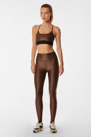 A woman stands against a plain background wearing a mid-tone brown gloss sports bra, matching Liquid Legging - Cafe high-waisted leggings made of Brazilian fabric, and beige sneakers with black accents. She has her hair tied back in a ponytail and bears a thoughtful expression.