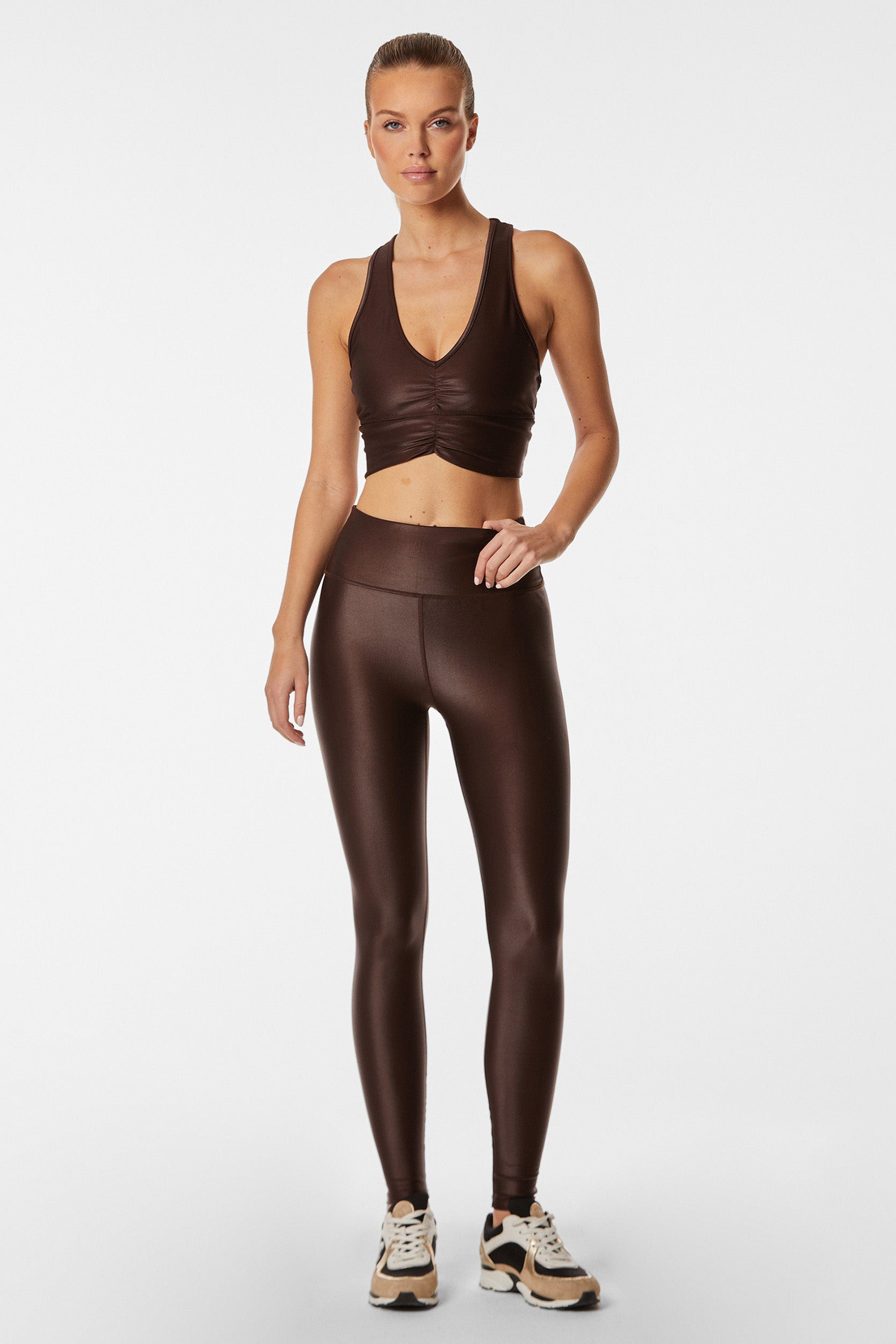 A person standing against a plain white background is wearing an espresso brown sports bra with a ruched front and matching Liquid Leggings - Espresso. They have on black, white, and beige sneakers. Their hair is tied back in a sleek bun, and they have a neutral facial expression.