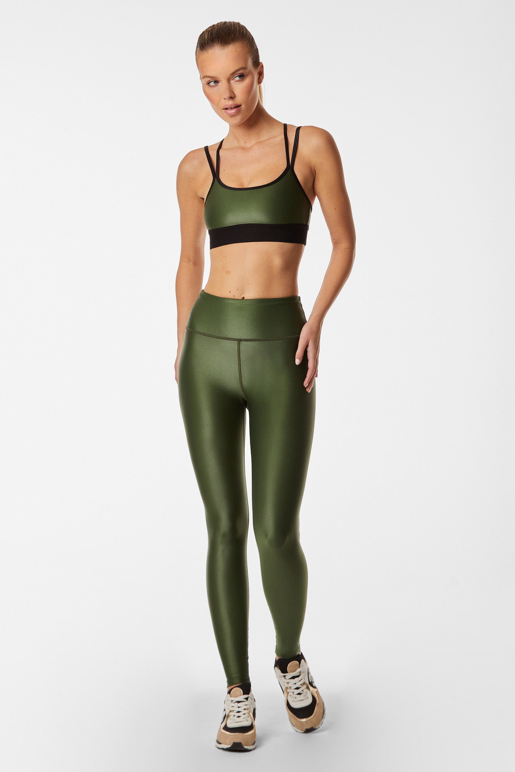 A person with light, tied-back hair is posing in a stylish ensemble featuring the Hunter Liquid Legging paired with a matching olive green sports bra accented by black straps. They are also wearing black and white sneakers. The background is plain white.