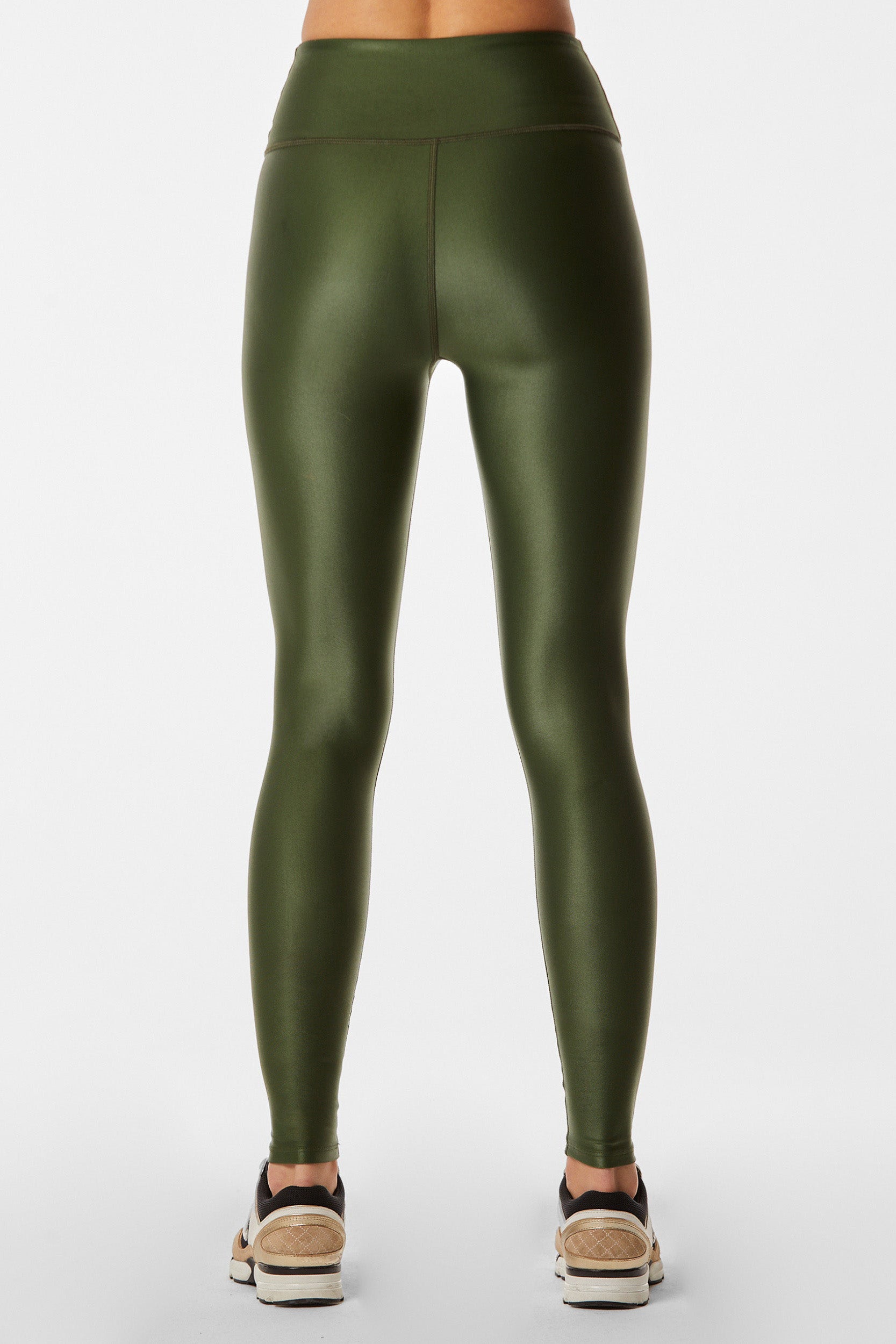 Back view of a person wearing the Liquid Legging - Hunter, high-waisted and squat-proof with a shiny dark green finish, paired with white sneakers featuring black and beige details. The background is plain white.