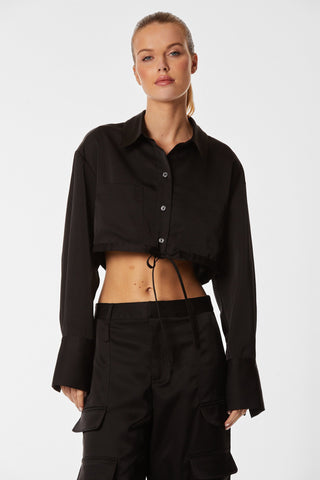 A person with long hair pulled back stands confidently, dressed in Noli Yoga's Milan Satin Button Up - Black, featuring long sleeves and extended cuffs. This luxurious shirt with a front pocket and buttons that reveal the midriff is paired with high-waisted tie-waist pants, creating a striking contrast against the plain white background.