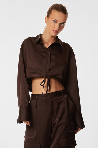 A woman with her hair tied back is wearing a dark brown, long-sleeved Milan Satin Button Up - Espresso shirt from Noli Yoga, along with a tie-waist and matching cargo pants featuring intricate cargo detailing. She is posing against a plain white background.