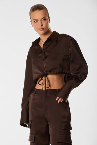 A woman wearing a dark brown Milan Satin Button Up - Espresso from Noli Yoga paired with matching cargo pants. Her blonde hair is styled in a sleek bun, and she gazes to the side with a neutral expression against a plain white background.