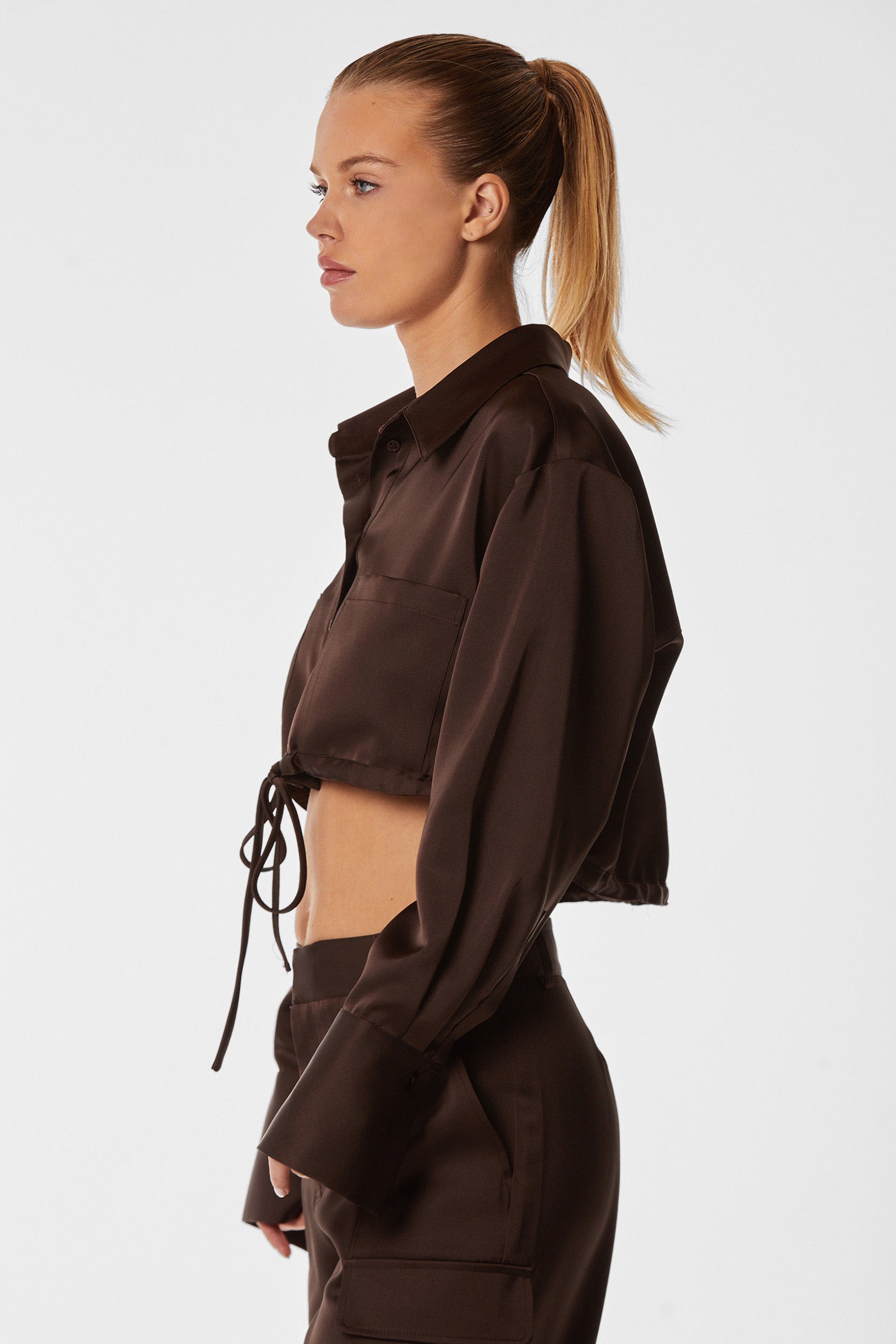 A woman with a ponytail is shown in profile wearing the Milan Satin Button Up - Espresso, a cropped, long-sleeve shirt with a tie-waist. She pairs it with matching high-waisted pants featuring cargo detailing and stands against a plain white background.