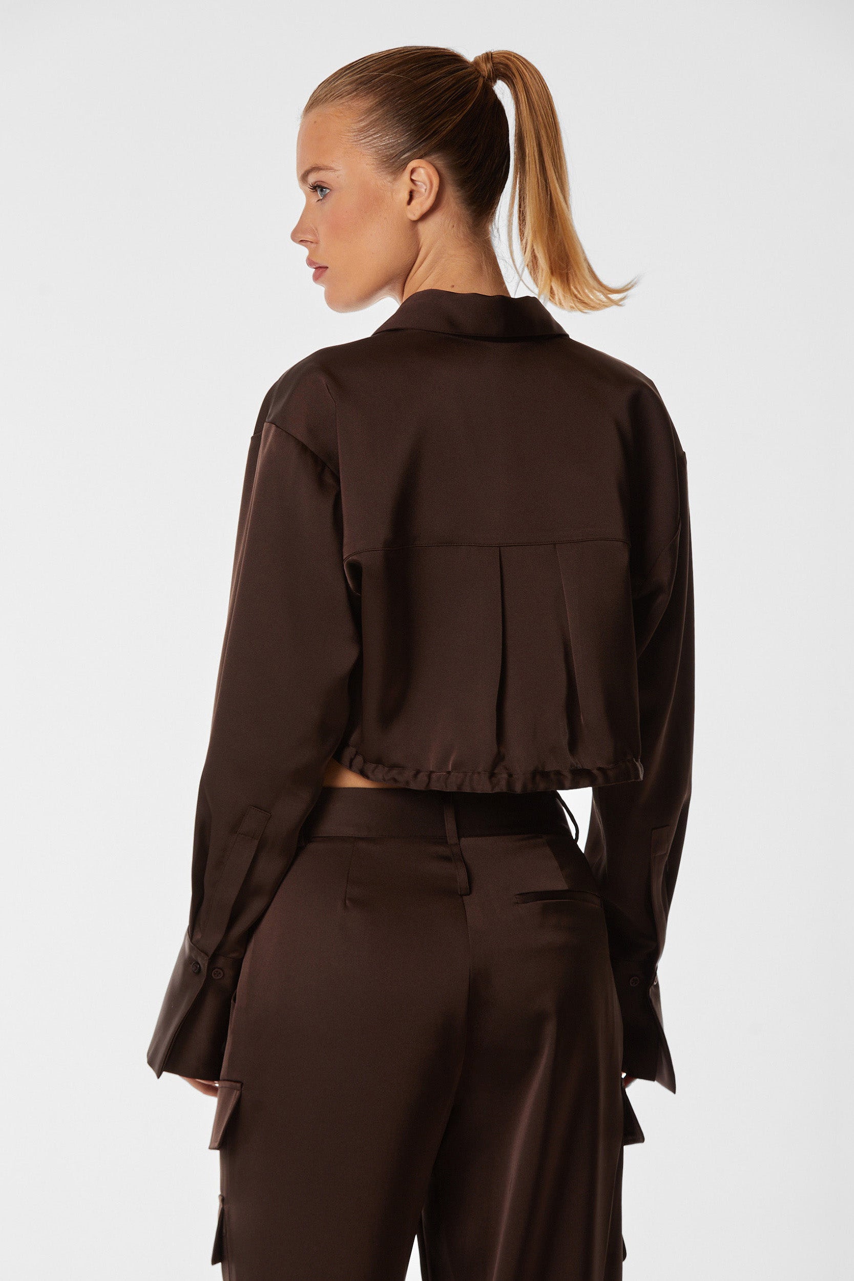 A woman with long blonde hair tied in a ponytail is shown from the side, wearing the Milan Satin Button Up in Espresso. The dark brown, long-sleeved cropped jacket and matching pants are made of smooth satin material with a slight sheen, featuring subtle cargo detailing for added style.