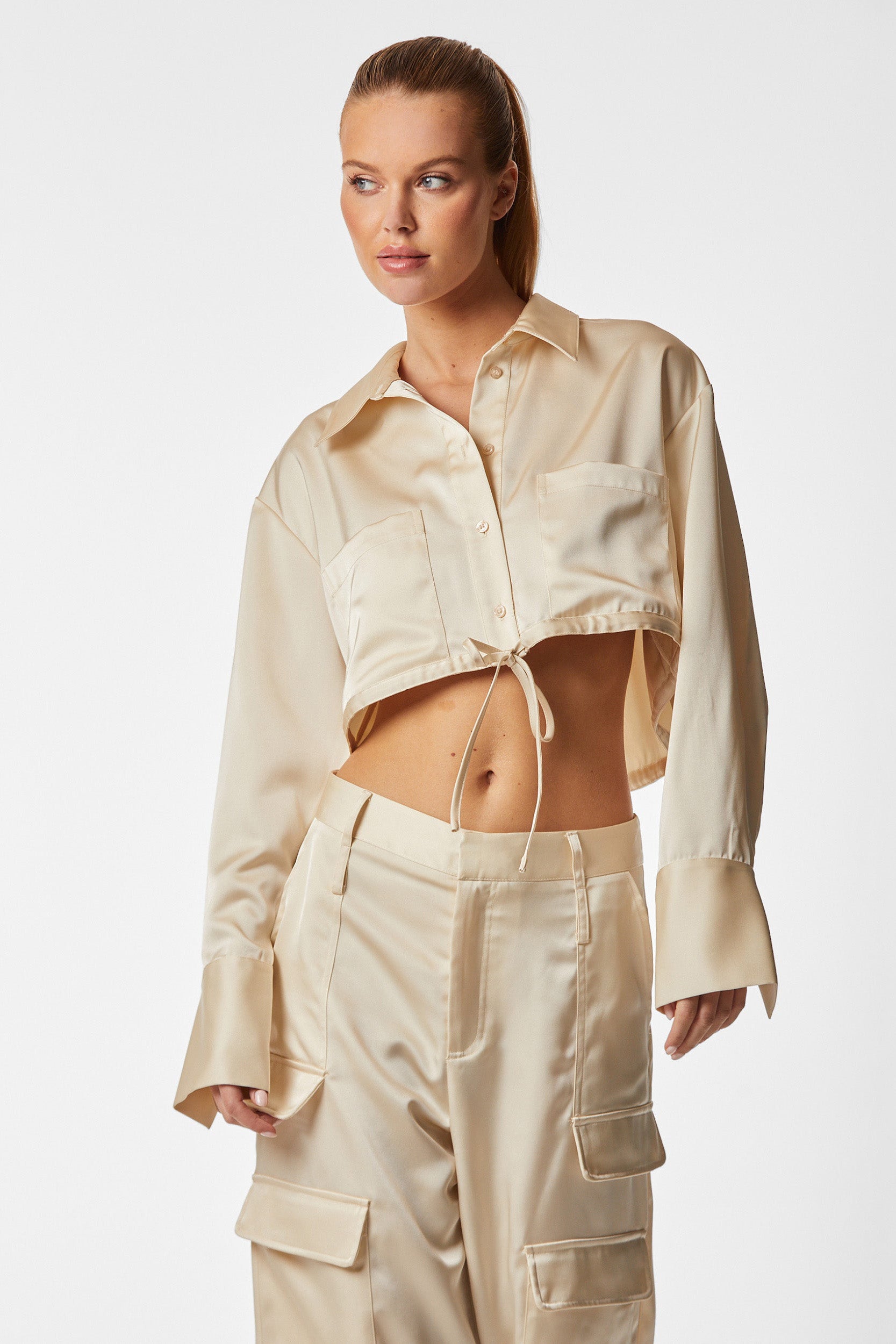 A woman is wearing a chic pearl-colored outfit featuring the Milan Satin Button Up - Pearl, a cropped satin shirt with an adjustable drawstring, complemented by matching high-waisted cargo pants adorned with intricate detailing. She has her hair tied back and poses against a plain white background, gazing slightly to the side.