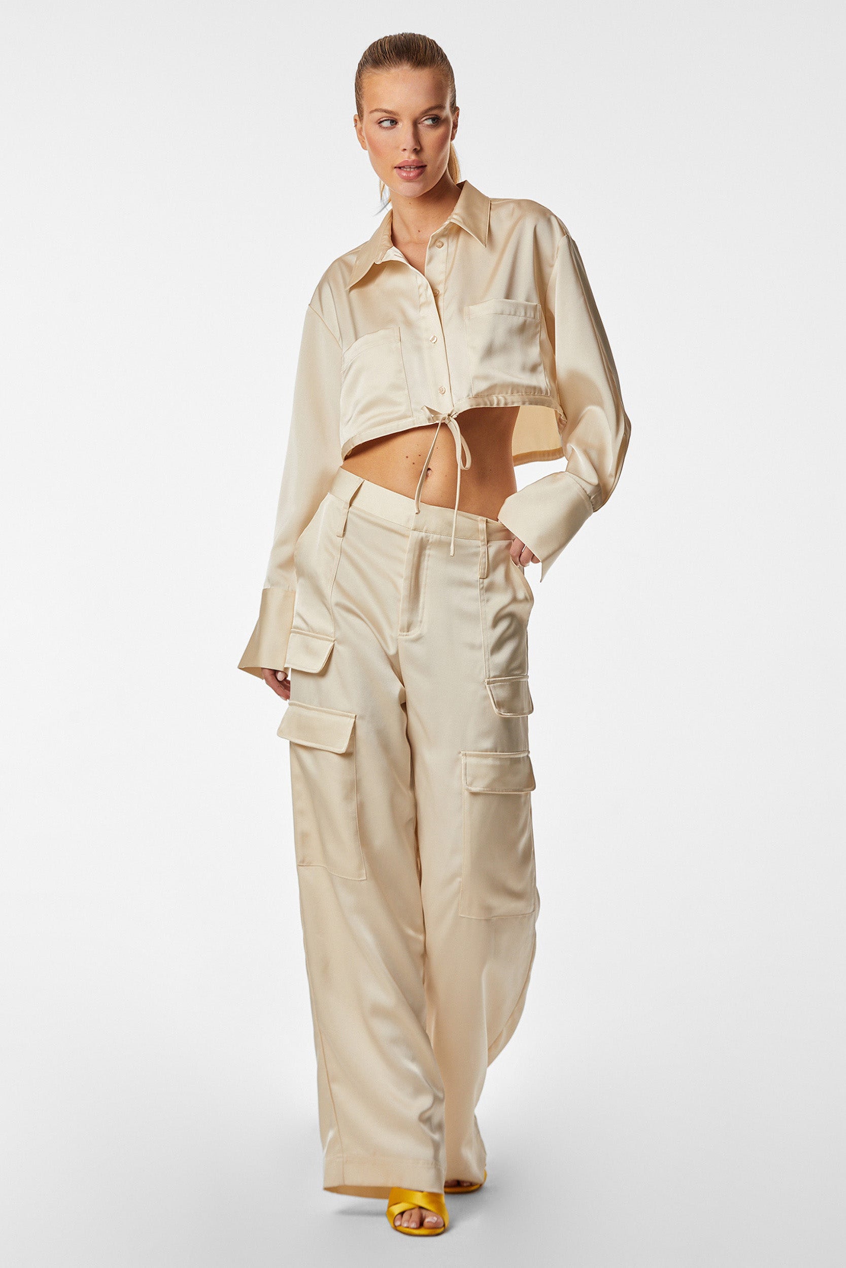 A woman models a stylish outfit featuring a cropped Pearl Milan Satin Button Up shirt tied at the waist and matching wide-leg cargo pants with multiple pockets. She wears yellow open-toe sandals and stands against a plain white background.