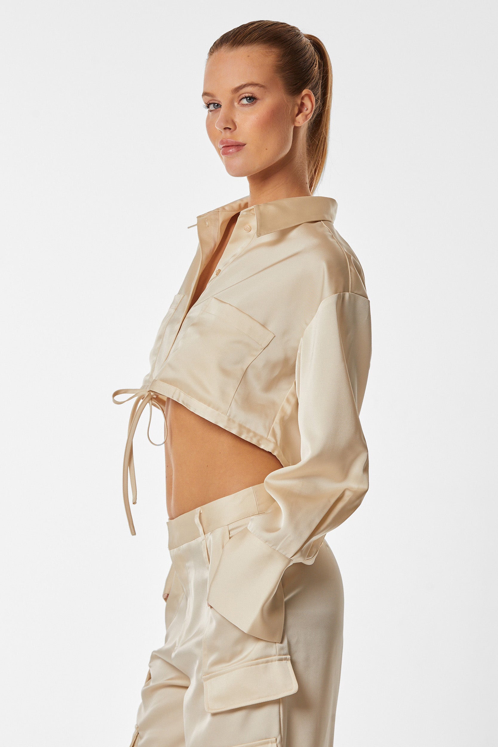 A person with a blonde ponytail wearing the Milan Satin Button Up - Pearl and matching pants featuring cargo detailing. They are standing sideways, looking toward the camera with a neutral expression against a plain white background.