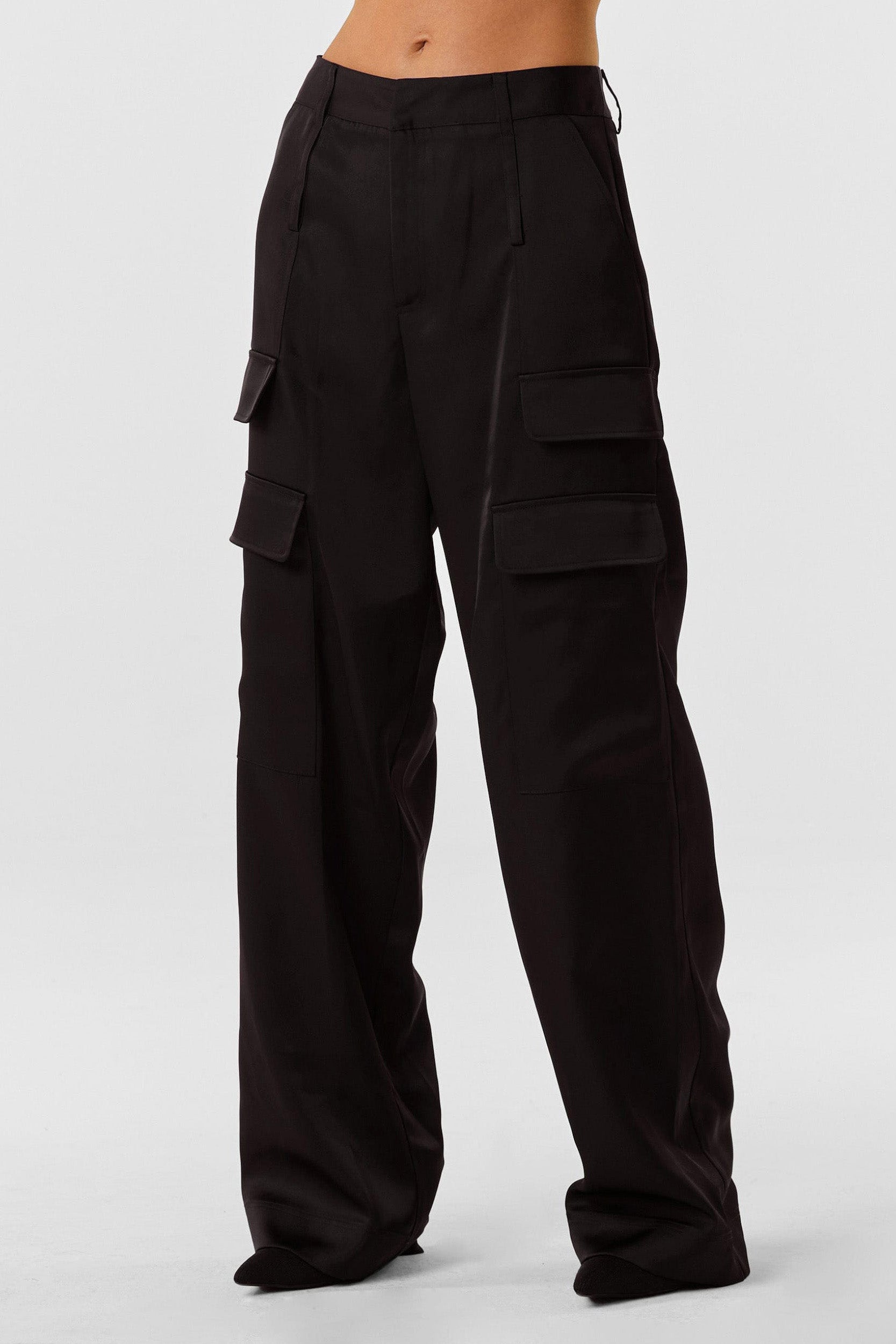 A person wearing the Milan Satin Cargo Pant - Black, featuring a high-waisted design and crafted from a satin-like fabric with multiple large pockets on the sides. The slightly loose-fitting pants extend down to cover the feet, and the plain white background emphasizes their unique mid-rise style.